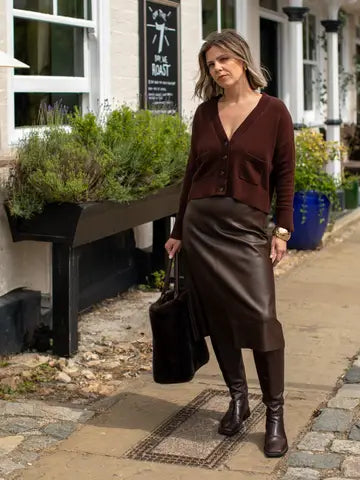 A woman stands on a sidewalk in front of a building, wearing a dark brown button-front cardigan, a brown faux-leather midi skirt, brown leggings, and brown knee-high boots, while holding a dark brown tote bag.