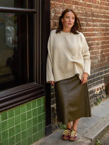 A woman stands outdoors against a brick wall and green tiled window, wearing an oversized cream knit sweater, a dark olive skirt, and brown sneakers.