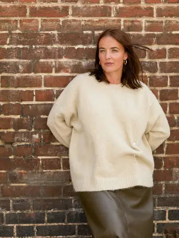 A woman wearing an oversized cream-colored knit sweater and dark brown leather pants, standing against a rustic red-brown brick wall.