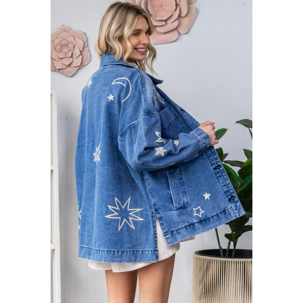 Woman wearing a blue denim jacket with white star and moon patterns, standing against a light-colored wall.