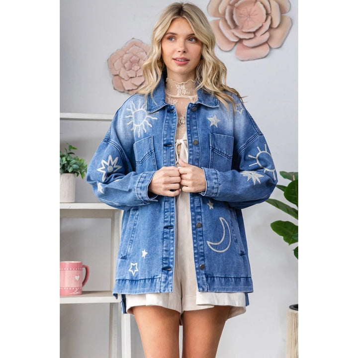 Woman wearing a blue denim jacket with white embroidery in a room setting.