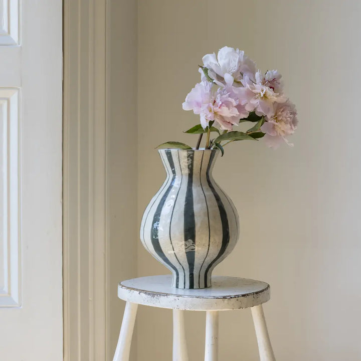 A ribbed ceramic vase with vertical black and white stripes, holding a bouquet of light pink peonies, styled on a distressed white wooden stool against a neutral beige wall.