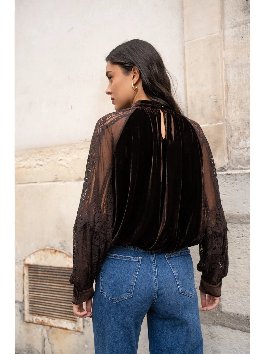Person wearing a sheer black blouse with cut-out shoulders and blue jeans against a stone wall.