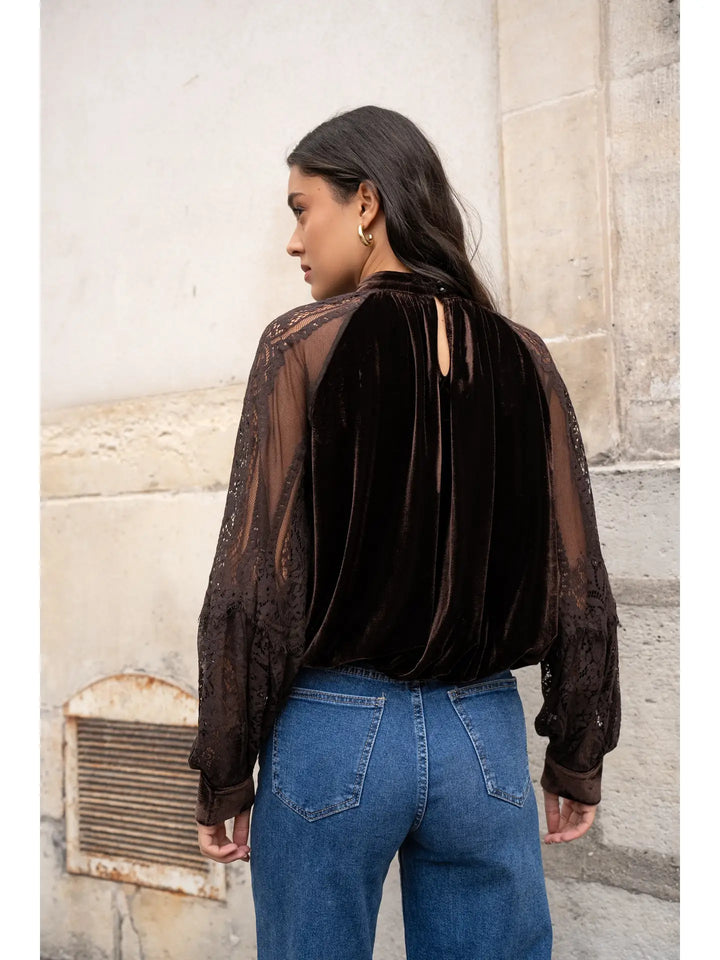 Person wearing a sheer black blouse with cut-out shoulders and blue jeans against a stone wall.