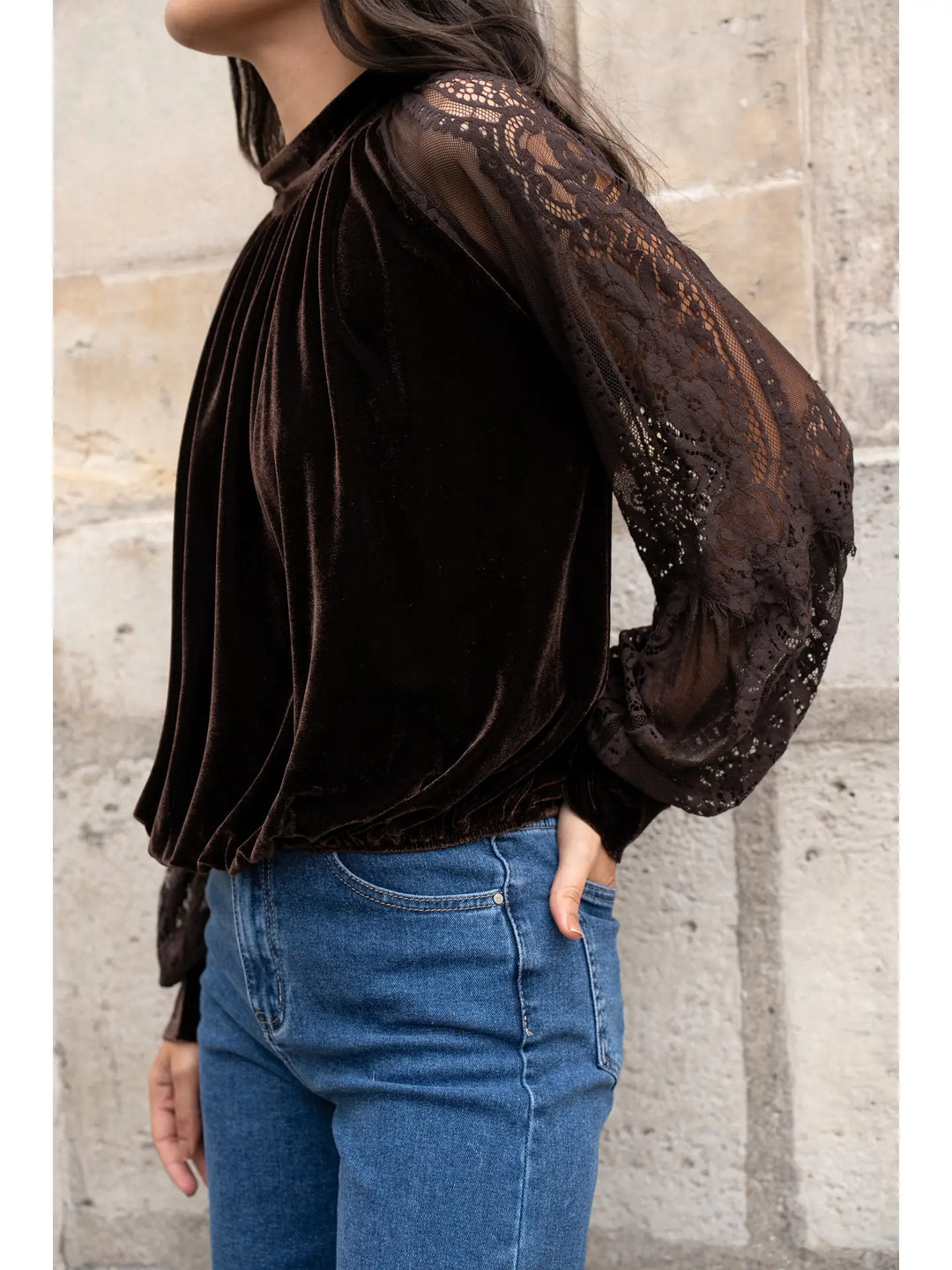 Person wearing a black velvet blouse with sheer lace sleeves against a stone wall.