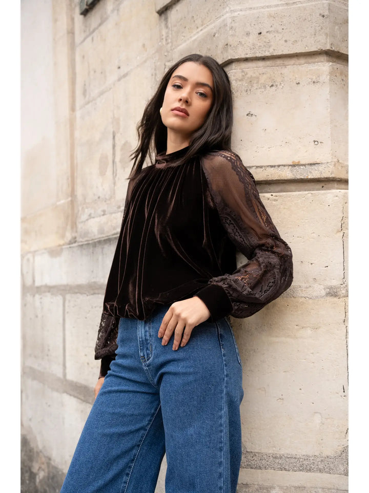 Woman wearing a dark blouse with sheer sleeves and blue jeans against a stone wall.