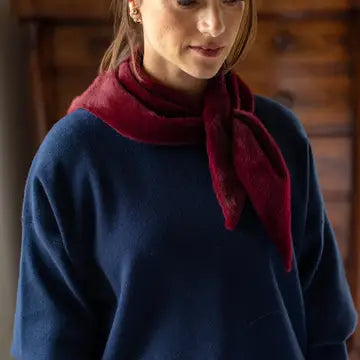 A woman wearing a navy blue knit sweater and a deep red scarf with a plush, velvety texture, photographed in a warm indoor setting with a wooden background.