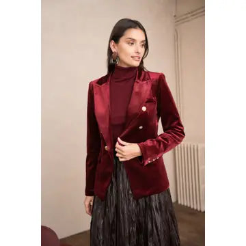 A woman wearing a deep burgundy velvet double-breasted blazer over a matching burgundy turtleneck sweater and a pleated black skirt, standing in an indoor setting.