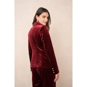 A woman wearing a deep burgundy velvet blazer and matching trousers, photographed from the back against a plain beige studio background.