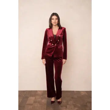 A woman modeling a deep burgundy velvet pantsuit, featuring a double-breasted blazer with gold buttons and matching wide-leg trousers, set against a neutral beige wall and wood floor.