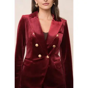 A woman wearing a deep burgundy velvet double-breasted blazer with gold buttons, shown from the chest up against a plain light background.