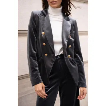 A woman wearing a dark gray velvet double-breasted blazer with gold buttons, styled with a white turtleneck and black trousers, set against a neutral stone wall background.