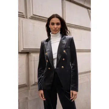 A woman stands outdoors against a light stone wall wearing a tailored double-breasted blazer in a dark navy velvet-like fabric with prominent gold buttons, paired with a white turtleneck and dark trousers.