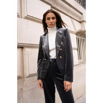 A woman stands outdoors in front of a light stone building, wearing a dark gray double-breasted velvet blazer with gold buttons over a white turtleneck and dark trousers.