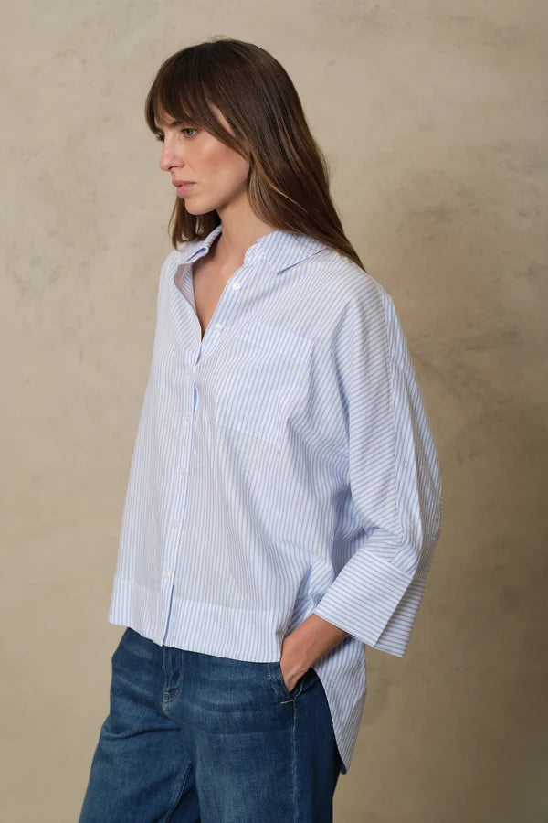 A woman wearing a light blue and white vertically striped button-up shirt with blue jeans, standing against a textured beige background.