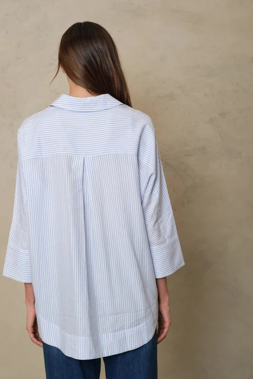 A woman viewed from behind wearing a light blue and white vertically striped button-up shirt with three-quarter sleeves and blue jeans, standing against a textured beige wall.