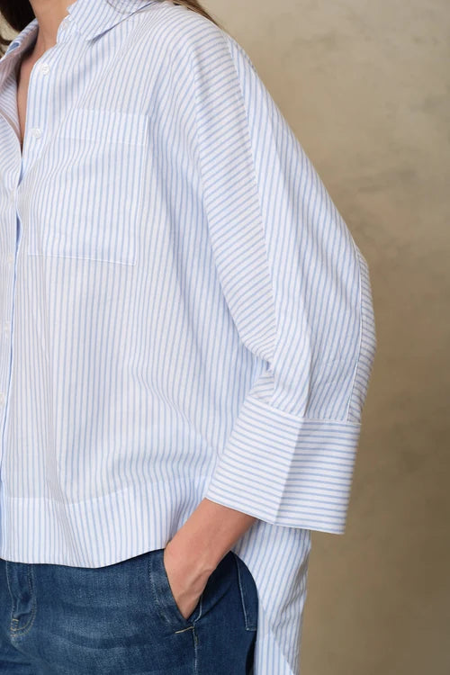 A close-up of a person wearing a white button-up shirt with thin vertical blue stripes, paired with dark blue jeans, against a neutral beige background.