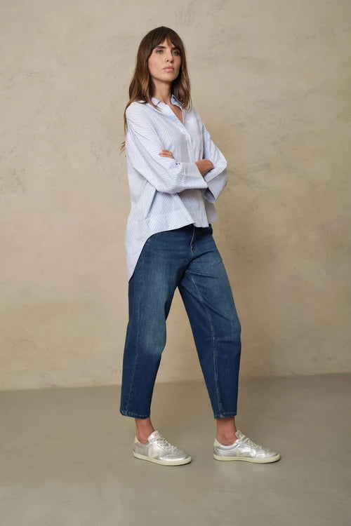 A woman stands with arms crossed wearing a light blue oversized button-up shirt, dark blue cropped jeans, and white sneakers against a textured beige studio backdrop.
