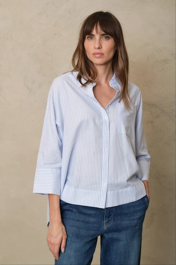 A woman wearing a light blue and white vertically striped button-down shirt with three-quarter sleeves, paired with blue jeans, standing against a textured beige wall.