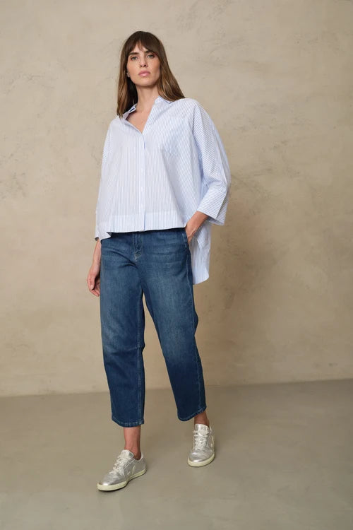 A woman modeling a light blue and white vertically striped button-down shirt with voluminous sleeves, paired with dark blue cropped jeans and white sneakers, standing against a textured beige studio wall.