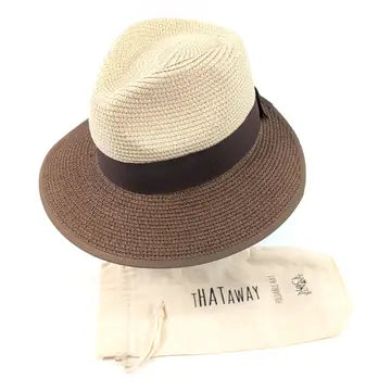 A woven fedora-style hat with a light beige crown, a dark brown band, and a medium brown brim, shown above a cream drawstring dust bag printed with the brand name THATAWAY.