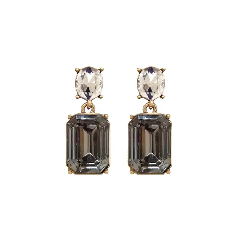 A pair of gold drop earrings featuring a large, faceted dark gray emerald-cut gemstone below a smaller, oval clear crystal, set against a plain white background.