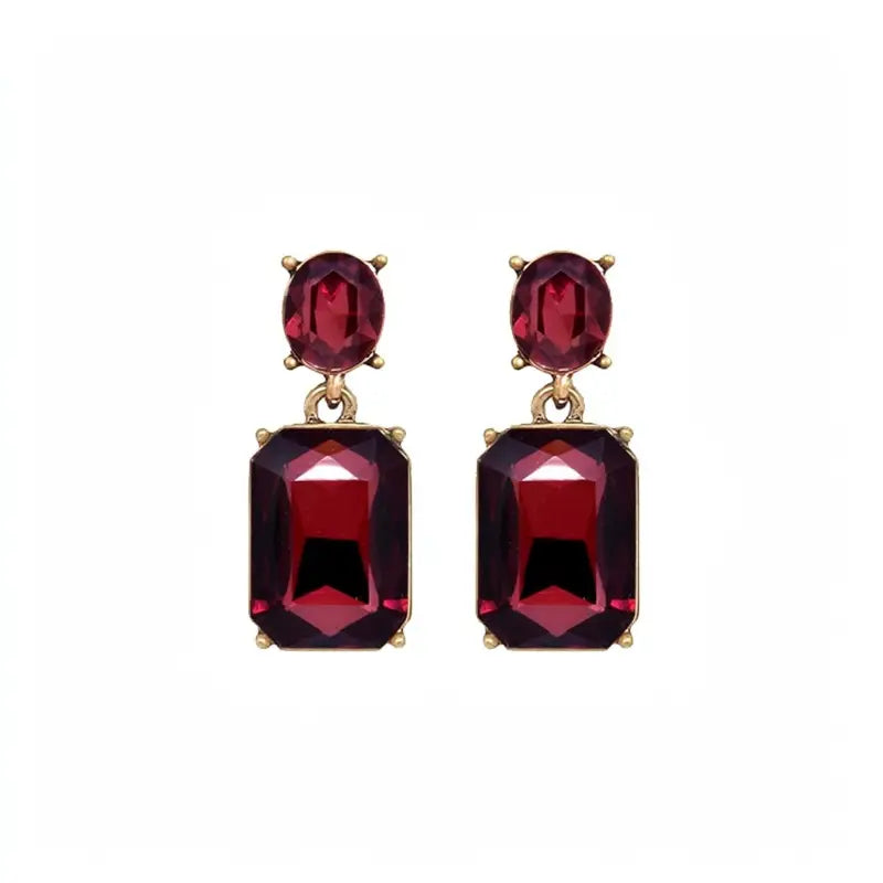 A pair of gold drop earrings featuring oval and rectangular deep red gemstones, set against a plain white background.