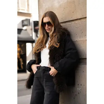 A woman wearing a black faux-fur trimmed puffer jacket, white top, and black jeans leans against a concrete wall in an outdoor urban setting.