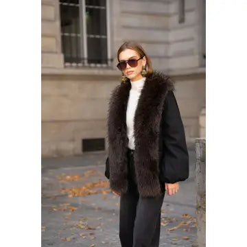 A woman wearing a black puffer jacket with a large dark brown fur collar, a white turtleneck, and black pants, standing outdoors in front of a stone building.