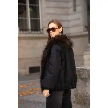 A woman wearing a black puffer jacket with a large dark brown fur collar and black sunglasses, standing outdoors in front of a stone building.