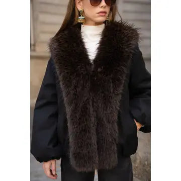 A woman wearing a black puffer jacket with a large, fluffy dark brown faux fur collar, styled with a white turtleneck, sunglasses, and statement earrings.