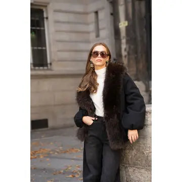 A woman wearing a black faux fur collar jacket over a white turtleneck and black trousers, standing outdoors against a stone building wall.