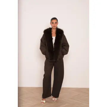 A model stands in a studio wearing a dark brown faux-fur trimmed jacket with matching wide-leg trousers, a white top, and white heeled sandals.