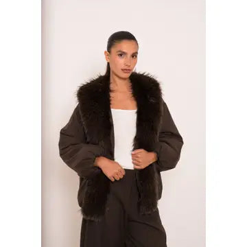 A model wearing a dark brown puffer jacket with a large dark brown faux fur collar, styled over a white top and dark pants, set against a plain off-white background.