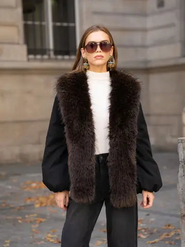 A woman wearing a black long-sleeve vest with a large dark brown fur collar, a white ribbed turtleneck, black pants, and sunglasses, standing outdoors in front of a stone building.