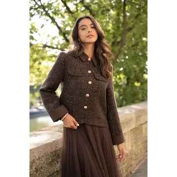 A woman wearing a brown tweed jacket with gold buttons and a matching brown pleated skirt, standing outdoors near a stone wall and green trees.