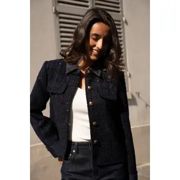 A woman wearing a dark navy textured tweed jacket with gold-tone buttons over a white top, standing in front of a gray shuttered window.