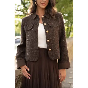 A woman wearing a cropped brown tweed jacket with gold-tone buttons over a white top and a dark brown pleated skirt, photographed outdoors with blurred greenery in the background.