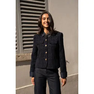 A woman wearing a dark navy textured tweed jacket with gold buttons and matching dark trousers, standing outdoors against a gray wall with a shuttered window.