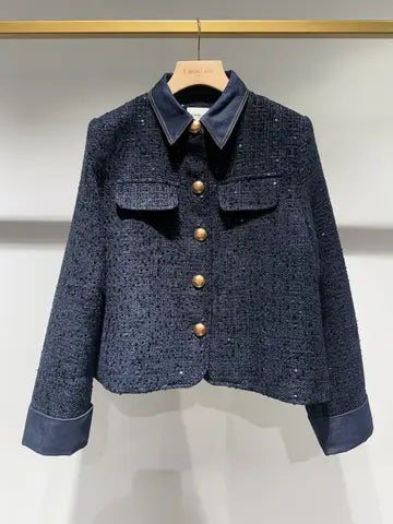 A dark navy blue tweed jacket with a contrasting solid navy collar, cuffs, and hem, featuring gold-tone buttons, hanging on a wooden hanger against a light gray wall.