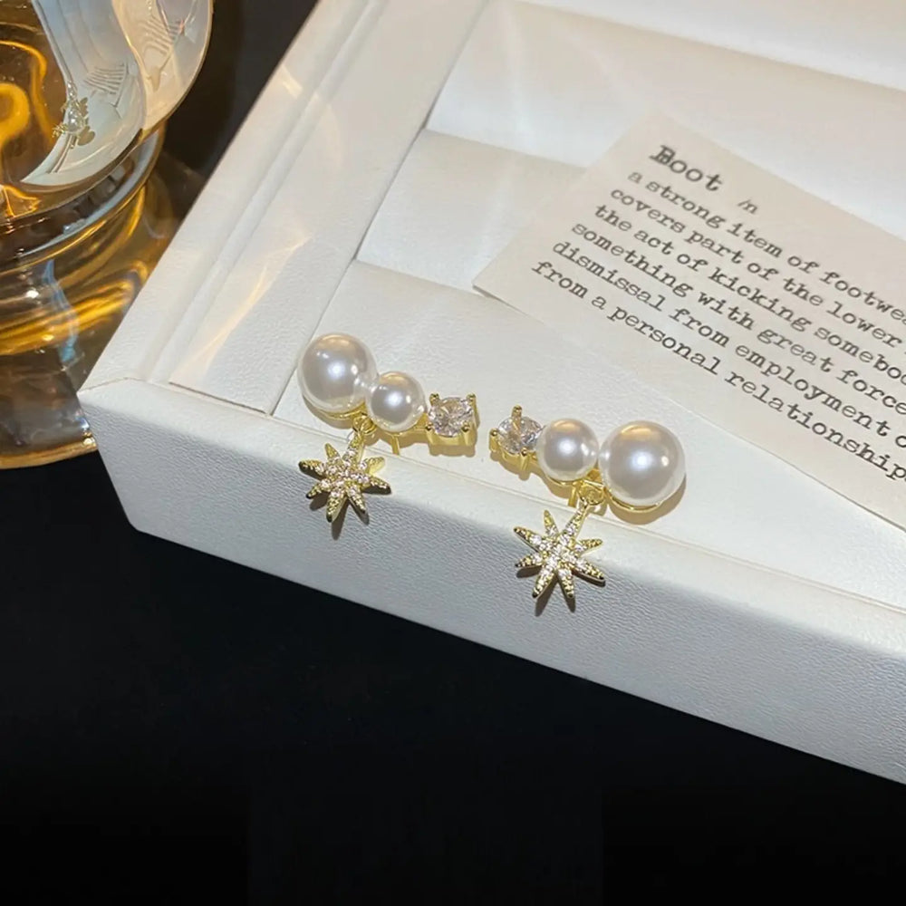 A pair of gold-toned stud earrings with white faux pearls, small clear crystal accents, and dangling starburst charms, displayed on a white jewelry box lid.