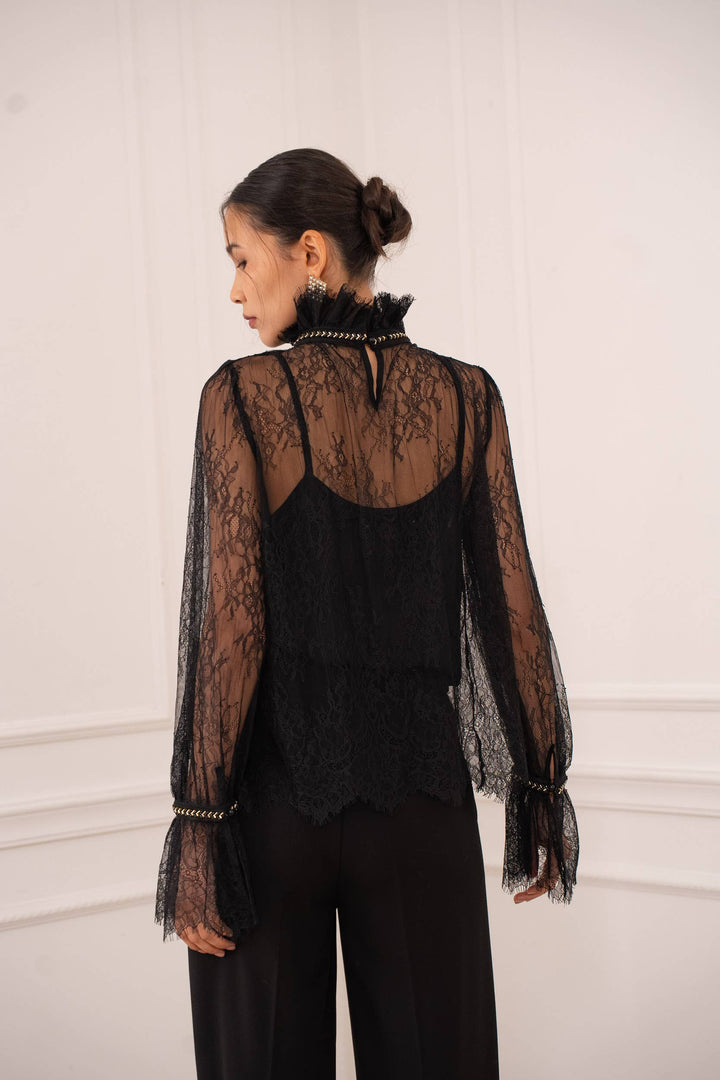 Person wearing a black lace top with sheer sleeves against a white wall.
