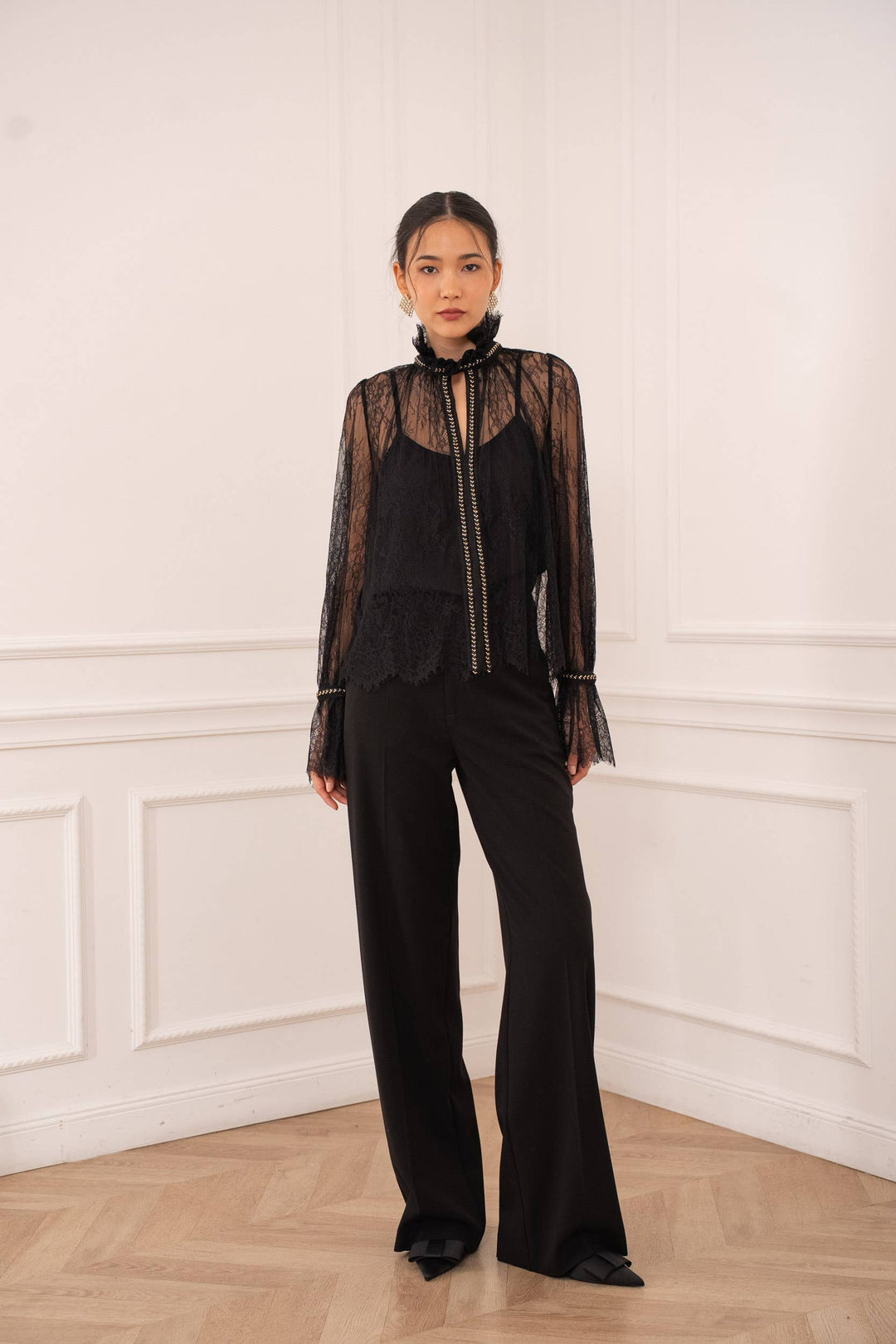 Woman wearing a black outfit with sheer overlay against a white wall.