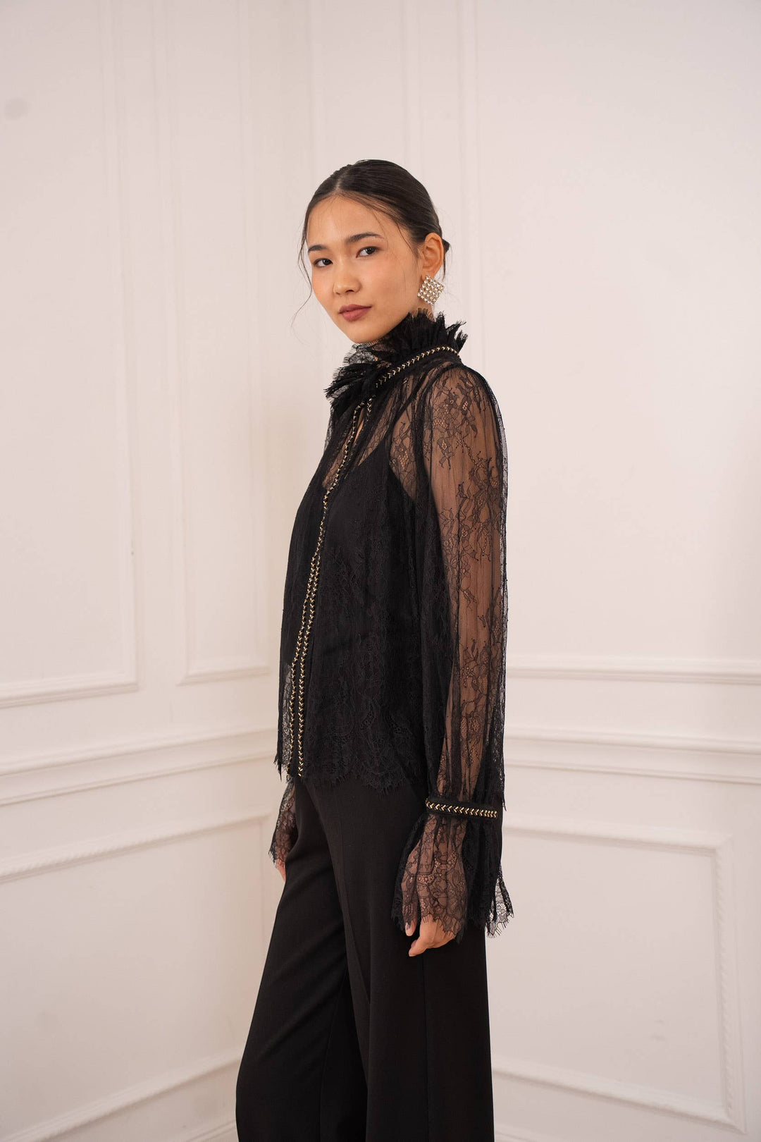Woman wearing a black lace top and black pants against a white wall