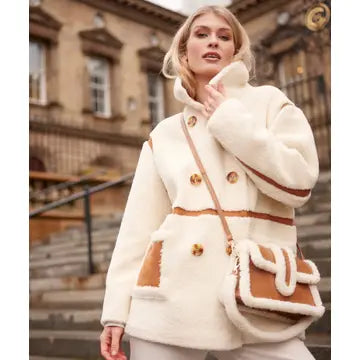 A woman wearing a cream-colored shearling-style coat with brown trim and gold buttons, carrying a matching cream and brown crossbody bag, standing outdoors in front of a stone building.