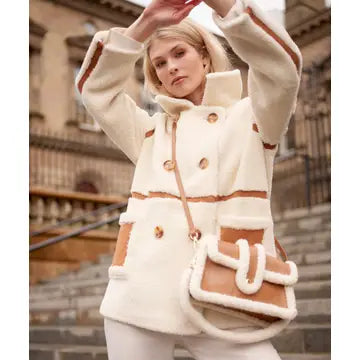 A woman wearing a cream-colored faux fur jacket with tan trim and buttons, paired with a matching cream and tan crossbody bag, set against a blurred background of stone steps and a building.