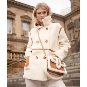 A woman wearing a cream-colored shearling-style coat with brown trim and buttons, carrying a matching cream and brown shoulder bag, standing outdoors in front of a stone building.