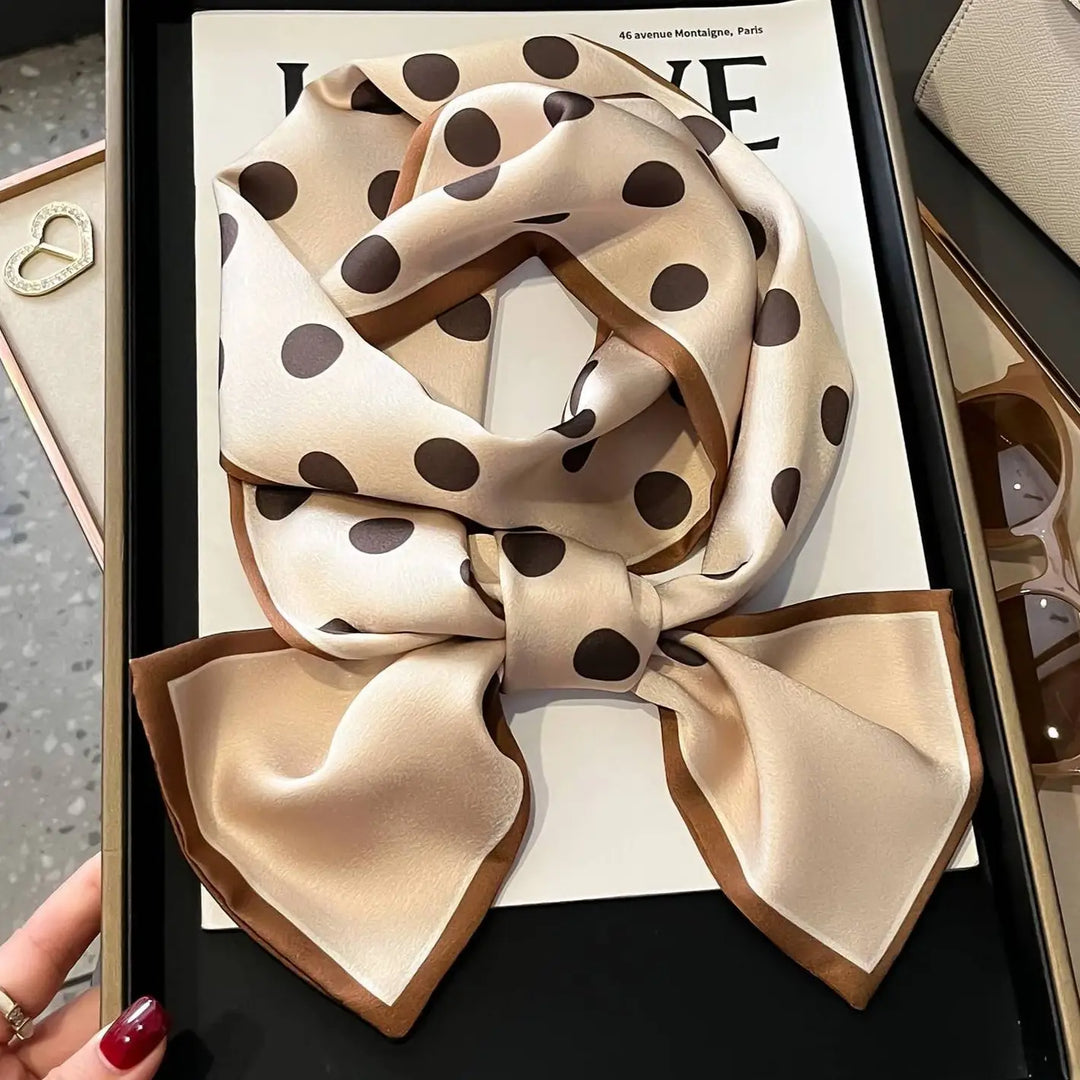 A beige silk scarf with dark brown polka dots and a thin brown border, styled in a large bow inside a black presentation box.