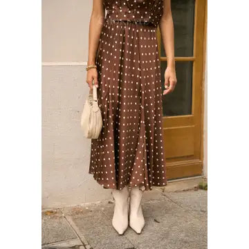 A woman wearing a brown midi dress with white polka dots, paired with white pointed-toe ankle boots and holding a cream-colored handbag, standing outdoors in front of a beige wall and wooden door.