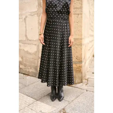 A sleeveless black midi dress with white polka dots, worn by a model standing outdoors against a stone wall.
